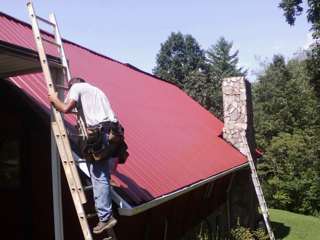 Roofer on a ladder beside a red metal roof and stone chimney — Soffit & Fascia Repair by Roofing Brothers in Blue Creek